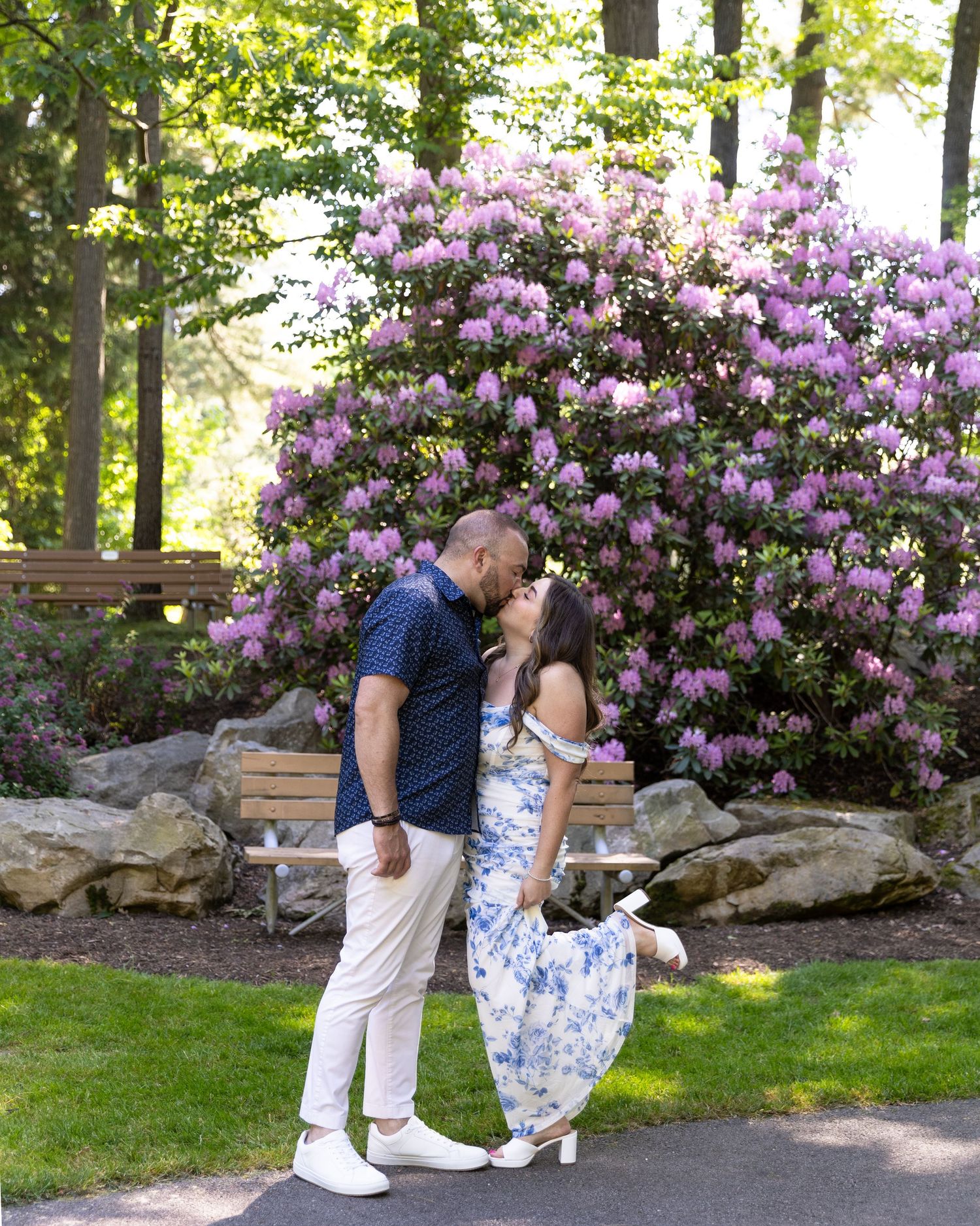 Couple sharing a romantic kiss in front of a blooming purple rhododendron bush in a lush green park.