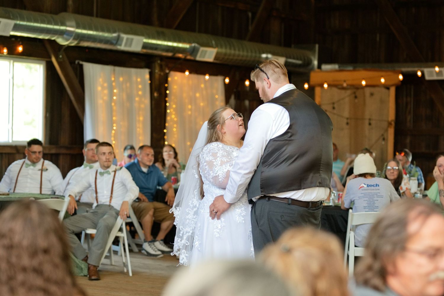 Bride and groom share first dance in rustic barn wedding venue with string lights and seated guests.