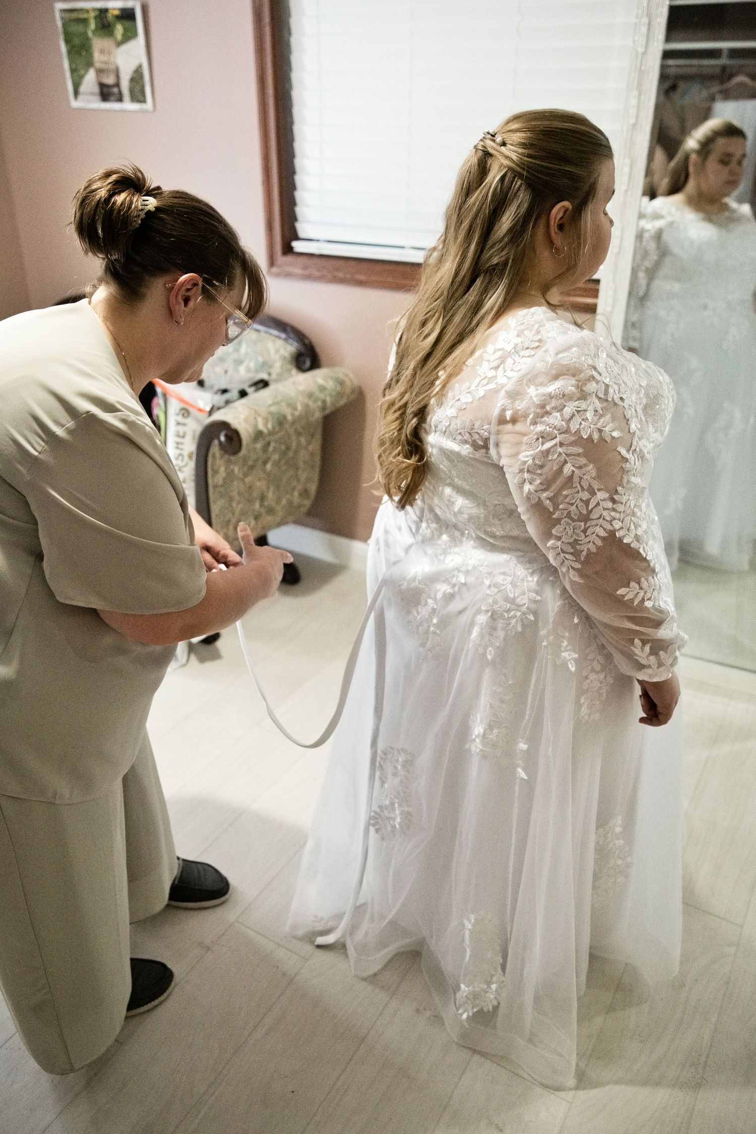A bridal consultant helping a bride in a lace wedding gown with long sleeves during a fitting in a bridal shop.