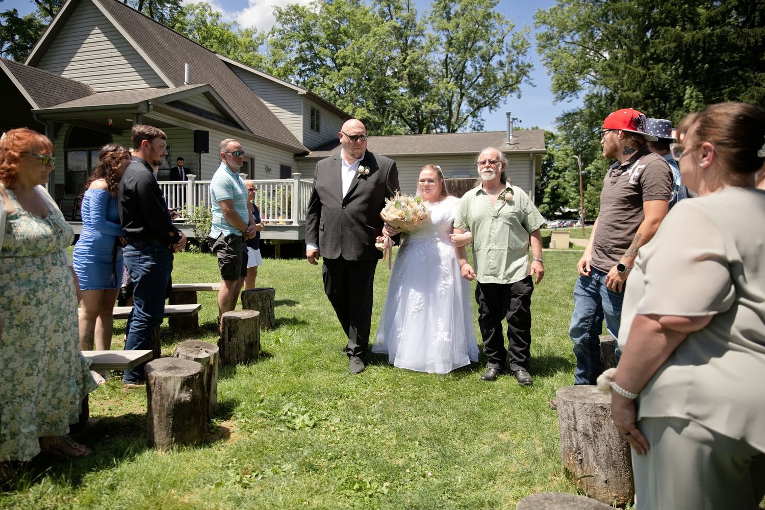 Bride in white gown walks down outdoor aisle with groom past tree stump seating at backyard wedding ceremony.