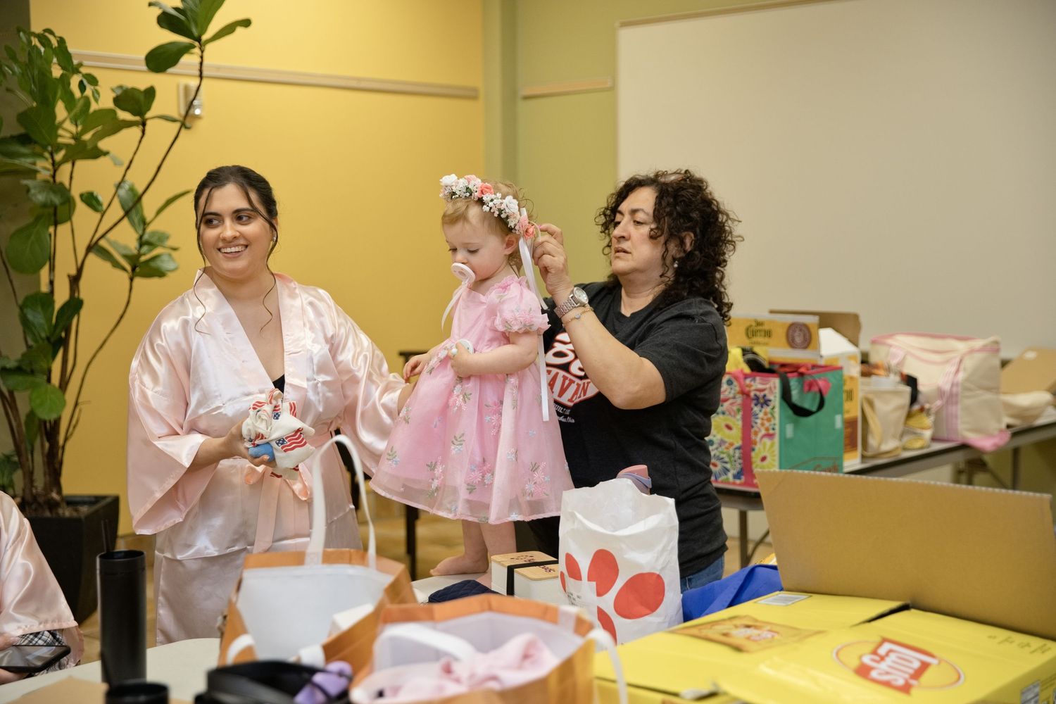 Two women and a toddler girl in pink dress with floral crown during a bridal preparation celebration.