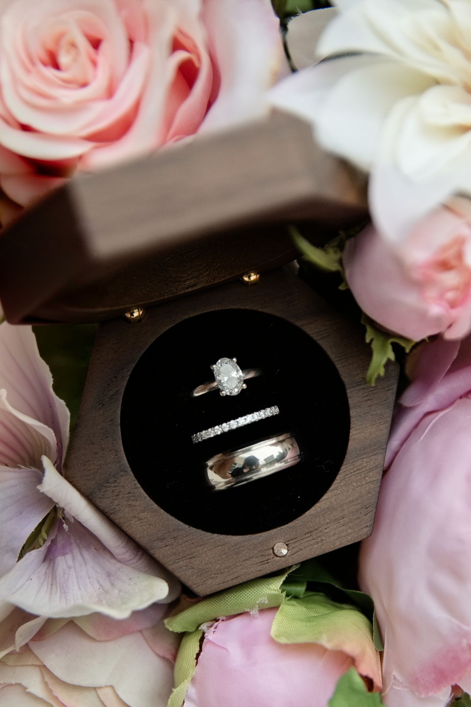Diamond engagement ring and wedding band displayed in a wooden ring box surrounded by pink roses and peonies.