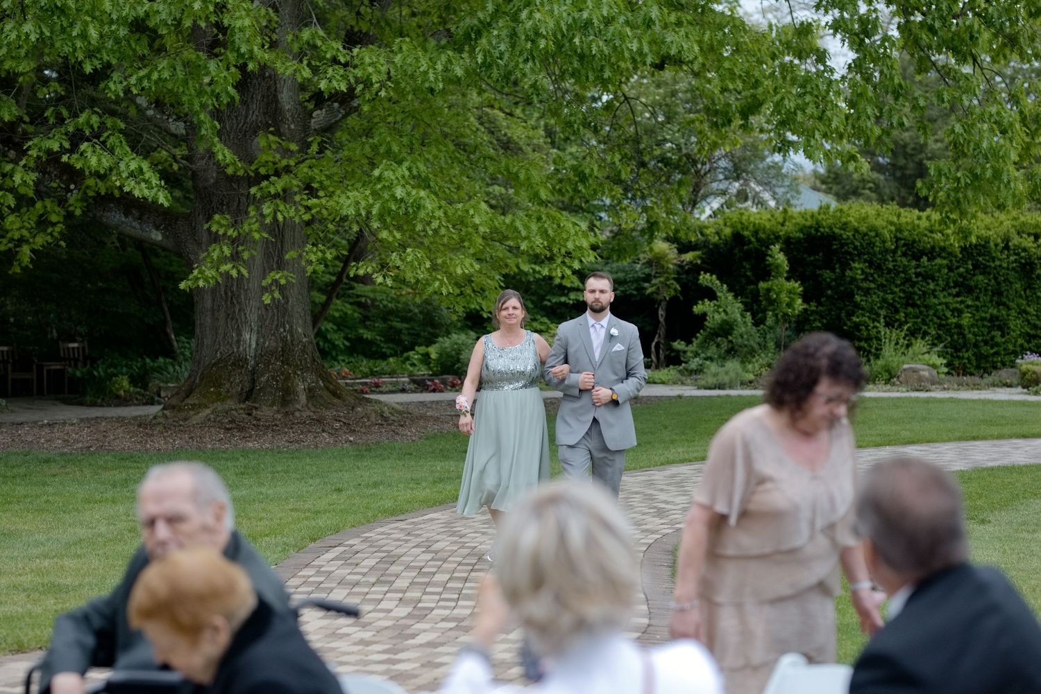 Couple walks arm-in-arm down a garden path at an outdoor wedding ceremony surrounded by lush green trees.