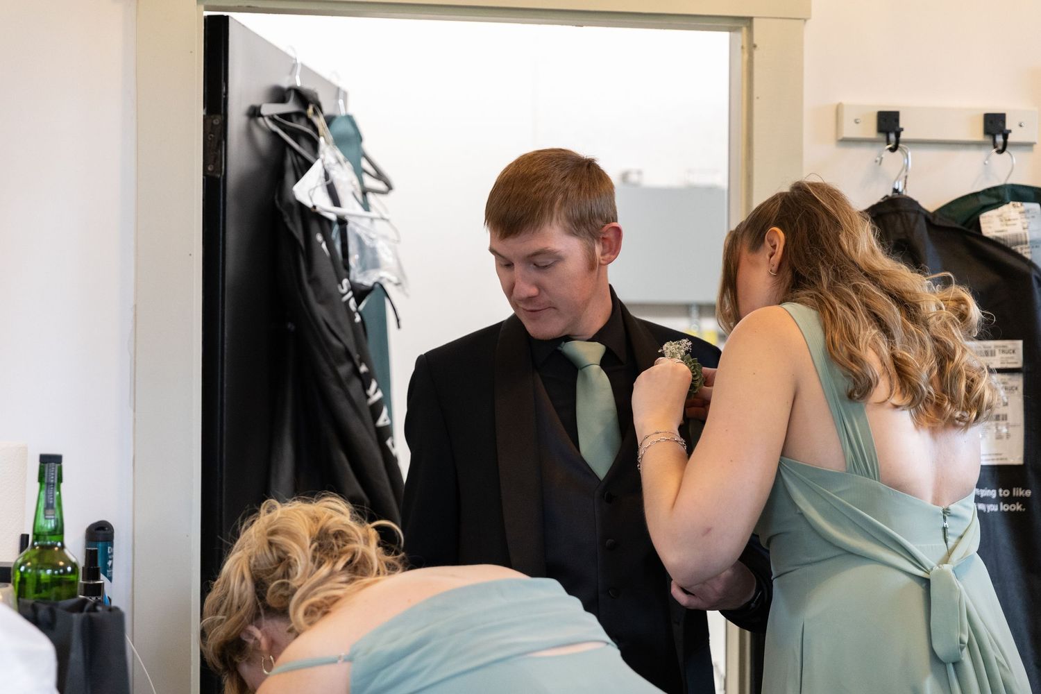 A young man in a black suit gets his boutonniere pinned by a woman in a sage green bridesmaid dress.