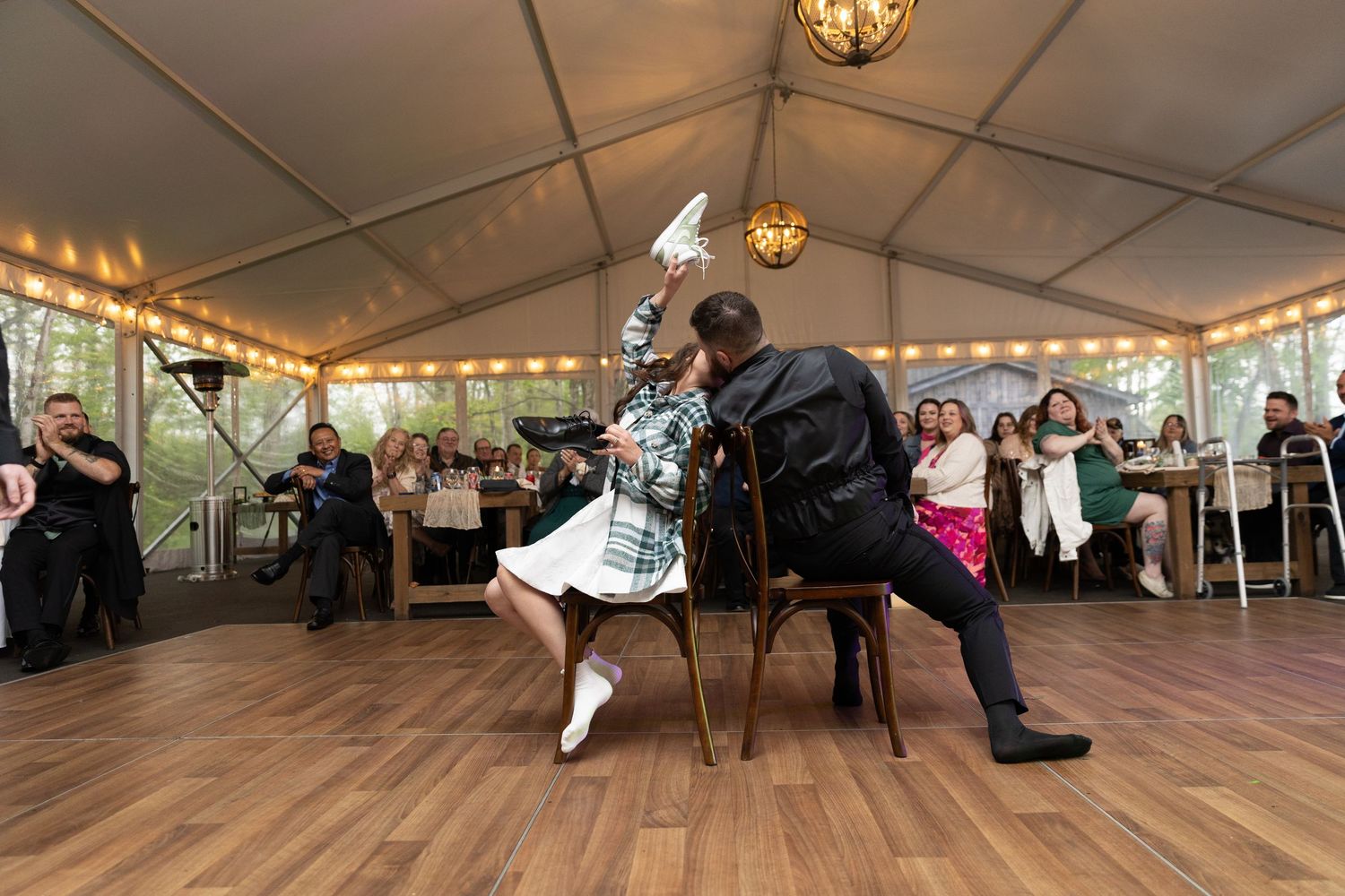 Couple plays musical chairs at an elegant outdoor wedding reception under a white tent with string lights.