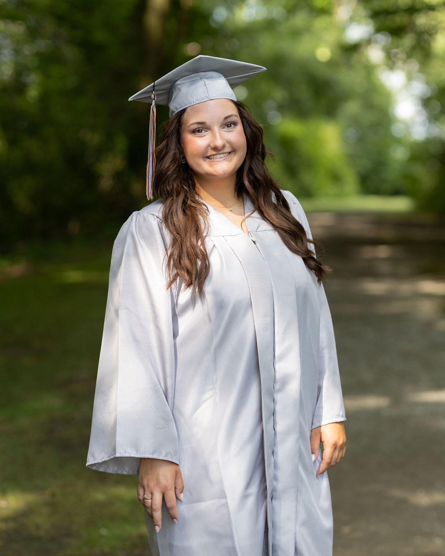 A graduate in white cap and gown poses outdoors on a wooded path.