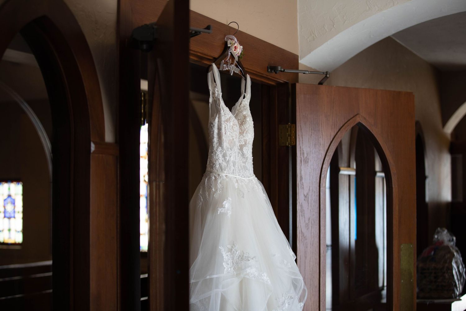 Elegant white lace wedding dress hanging in a church with arched wooden doorways.