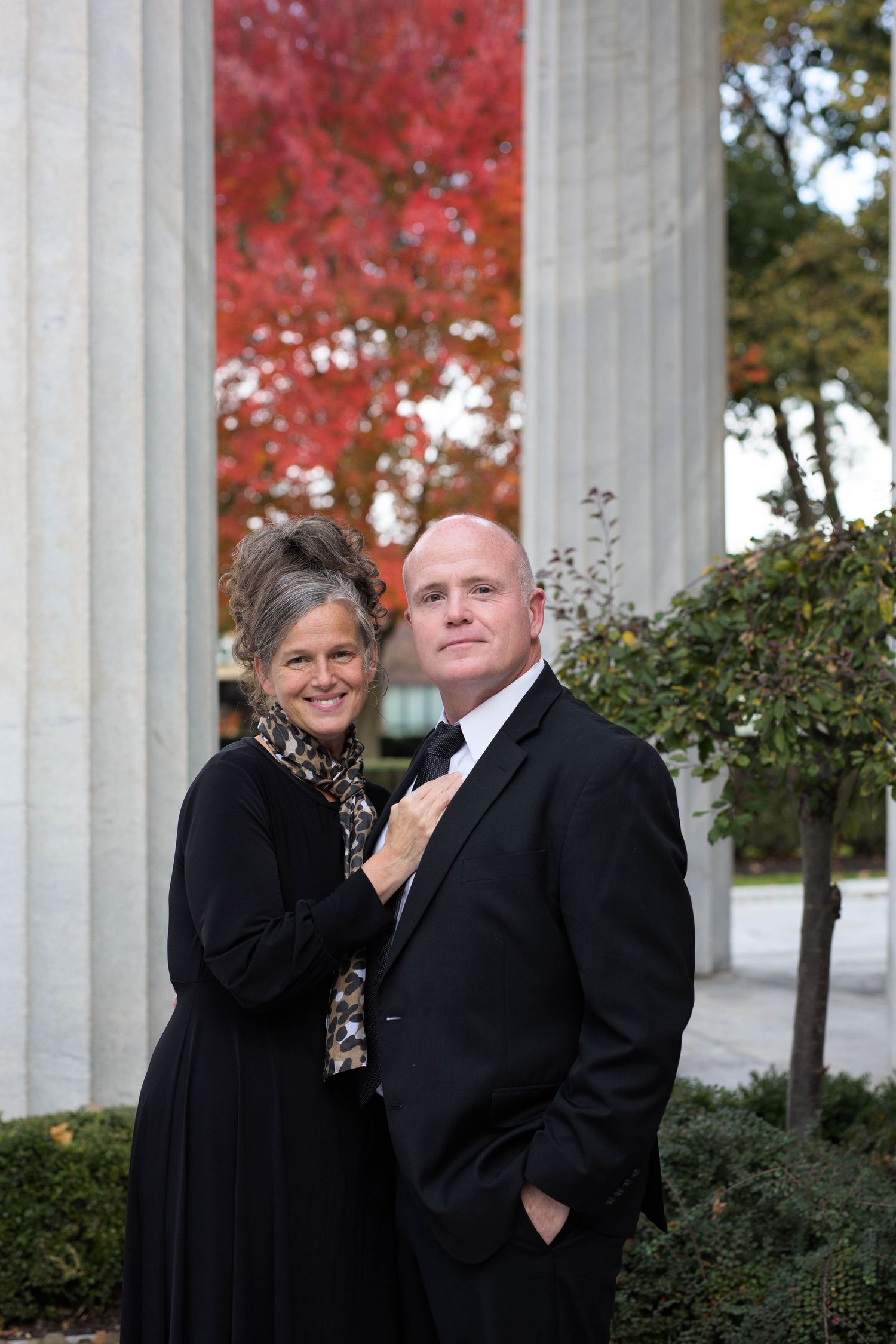 A formal couple poses between white columns with vibrant red fall foliage in background.