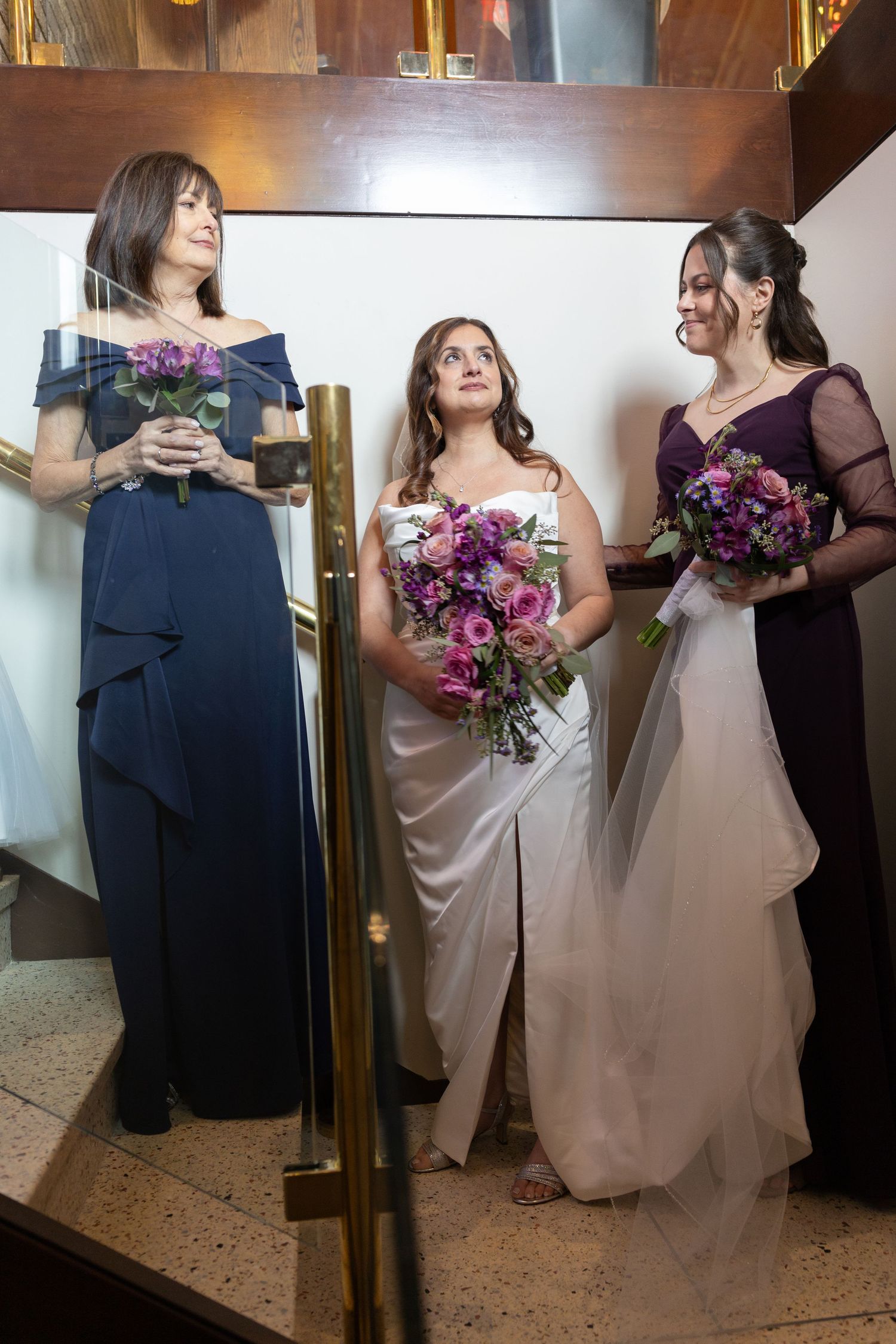 Wedding party members holding purple bouquets in a hallway.