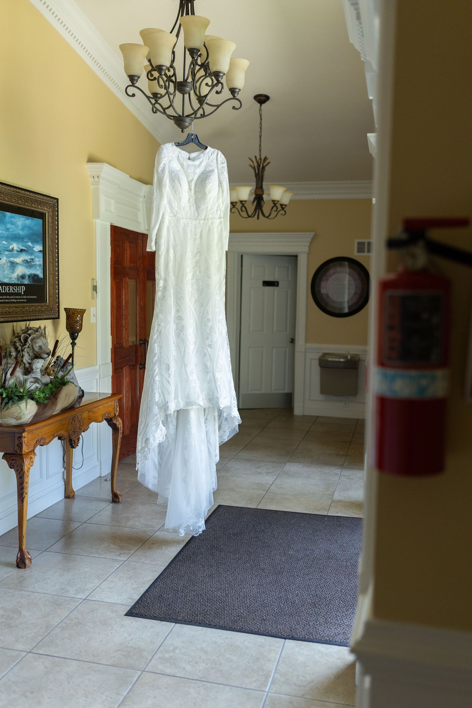 An elegant white wedding dress hangs from a chandelier in a foyer with yellow walls and tile floors.