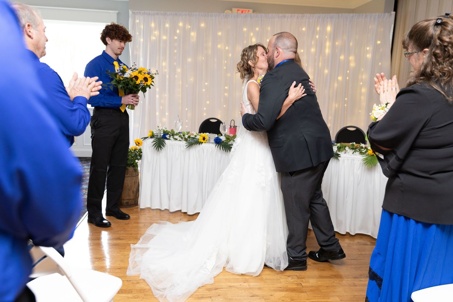 Bride and groom share first kiss at wedding reception while guests applaud, with sunflowers and fairy lights in background.