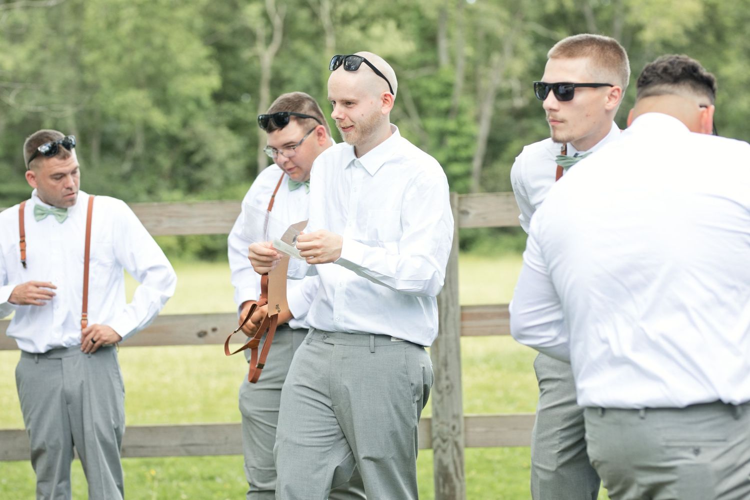 Groomsmen in white shirts and gray pants with suspenders gather outdoors near a wooden fence before a wedding ceremony.