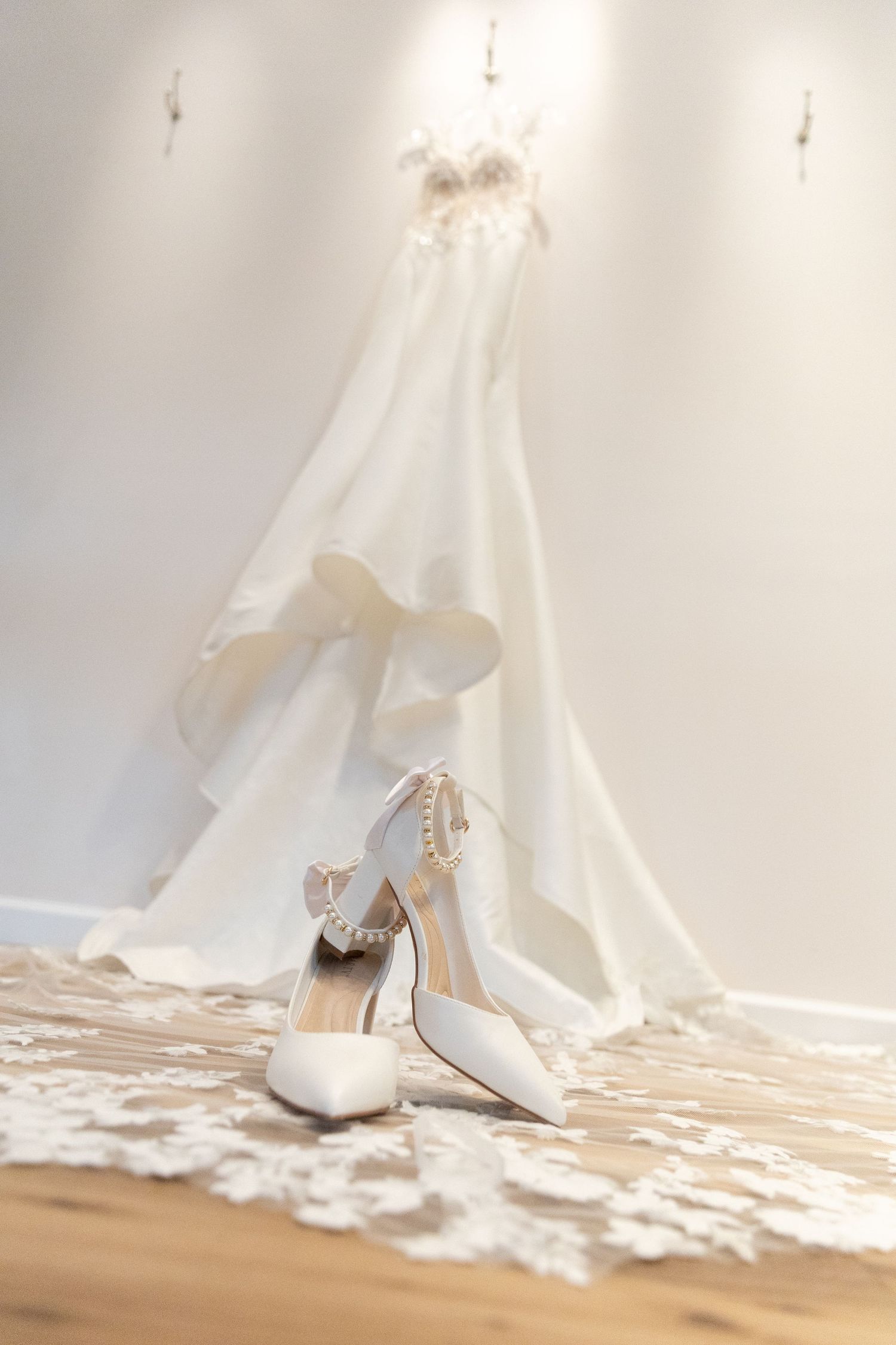 Elegant white bridal heels with pearl ankle straps displayed on wooden floor before a flowing wedding dress.