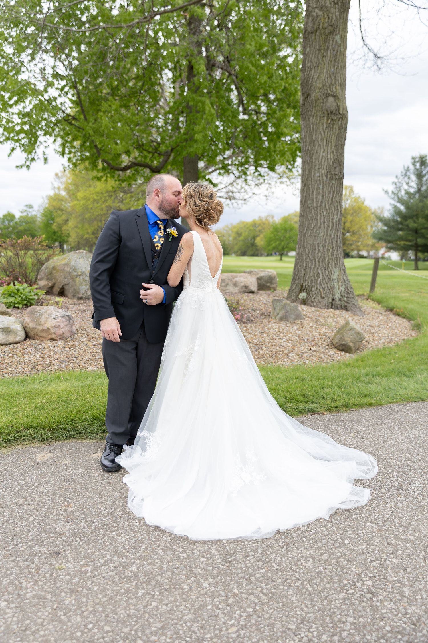 Bride and groom share a kiss outdoors, bride in elegant white gown with long train, groom in dark suit with blue tie.