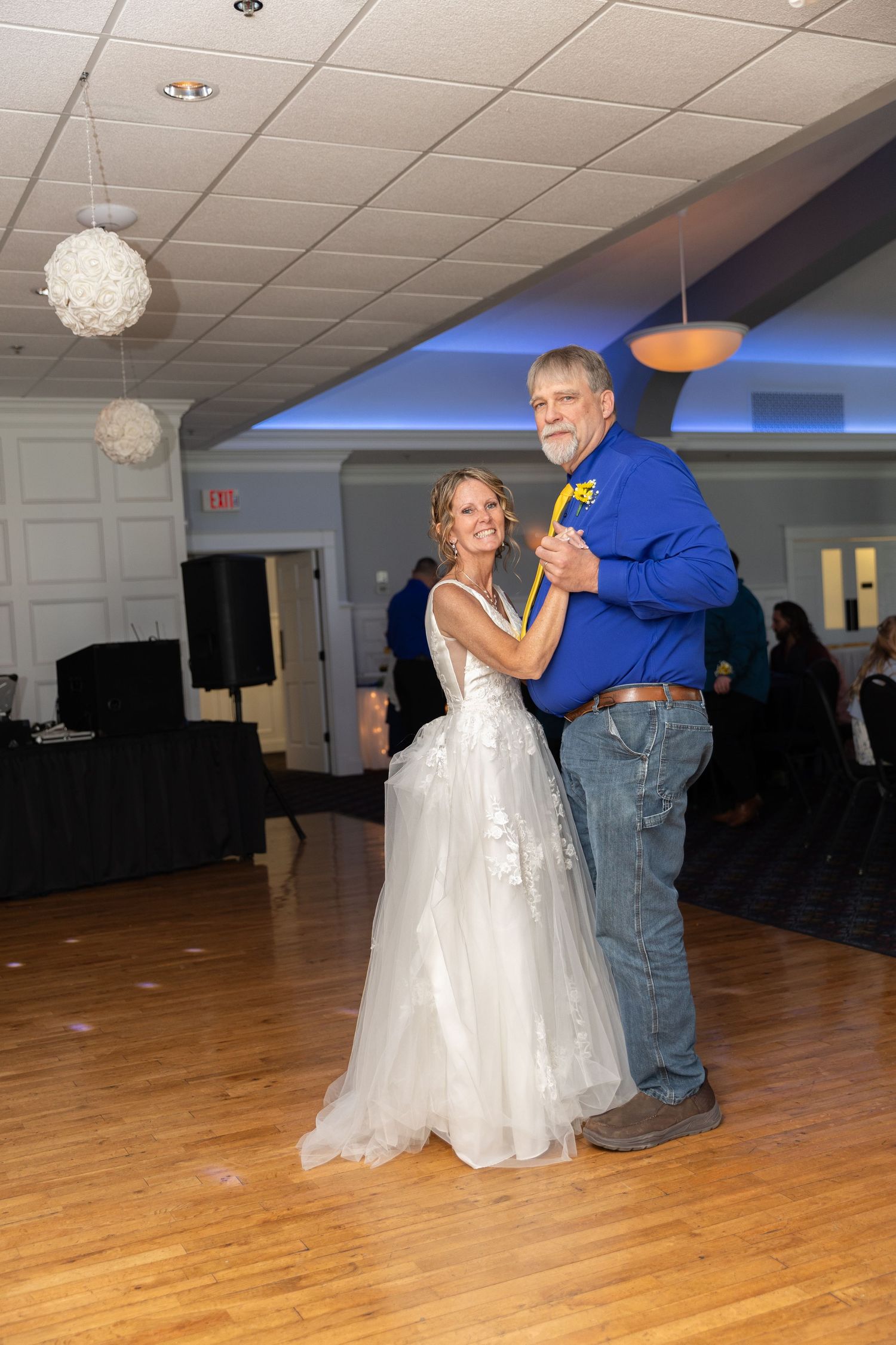 Bride in white gown shares father-daughter dance with man in blue shirt at elegant wedding reception.