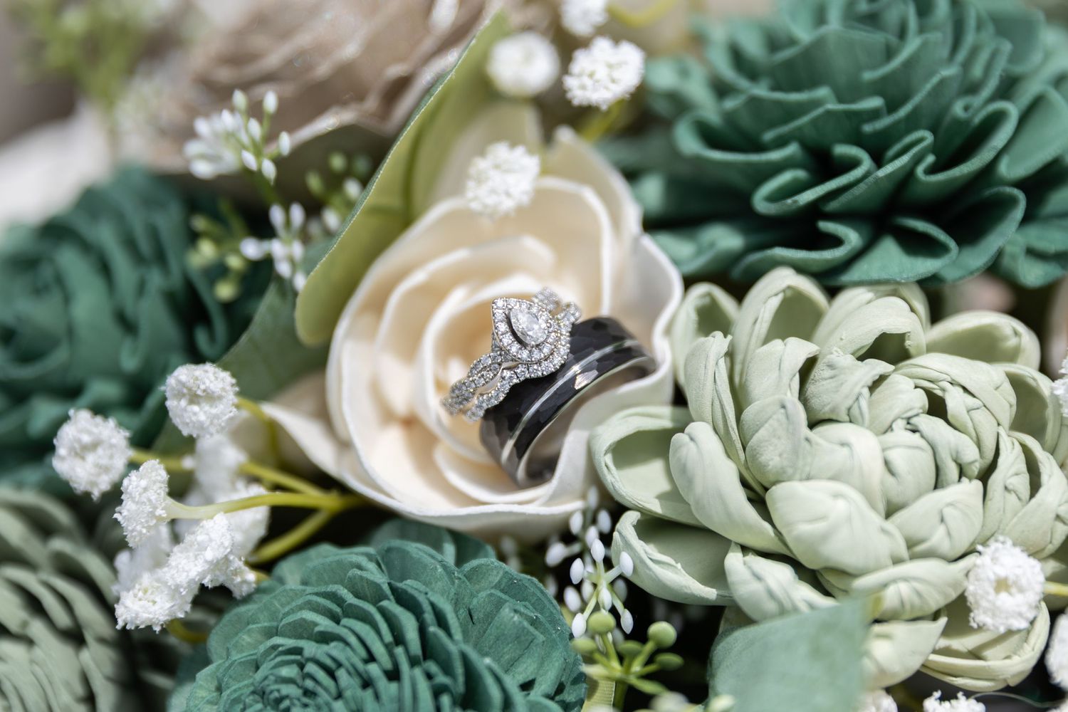 Wedding rings nestled among green and white sola wood flowers, featuring a diamond halo engagement ring and black wedding bands.