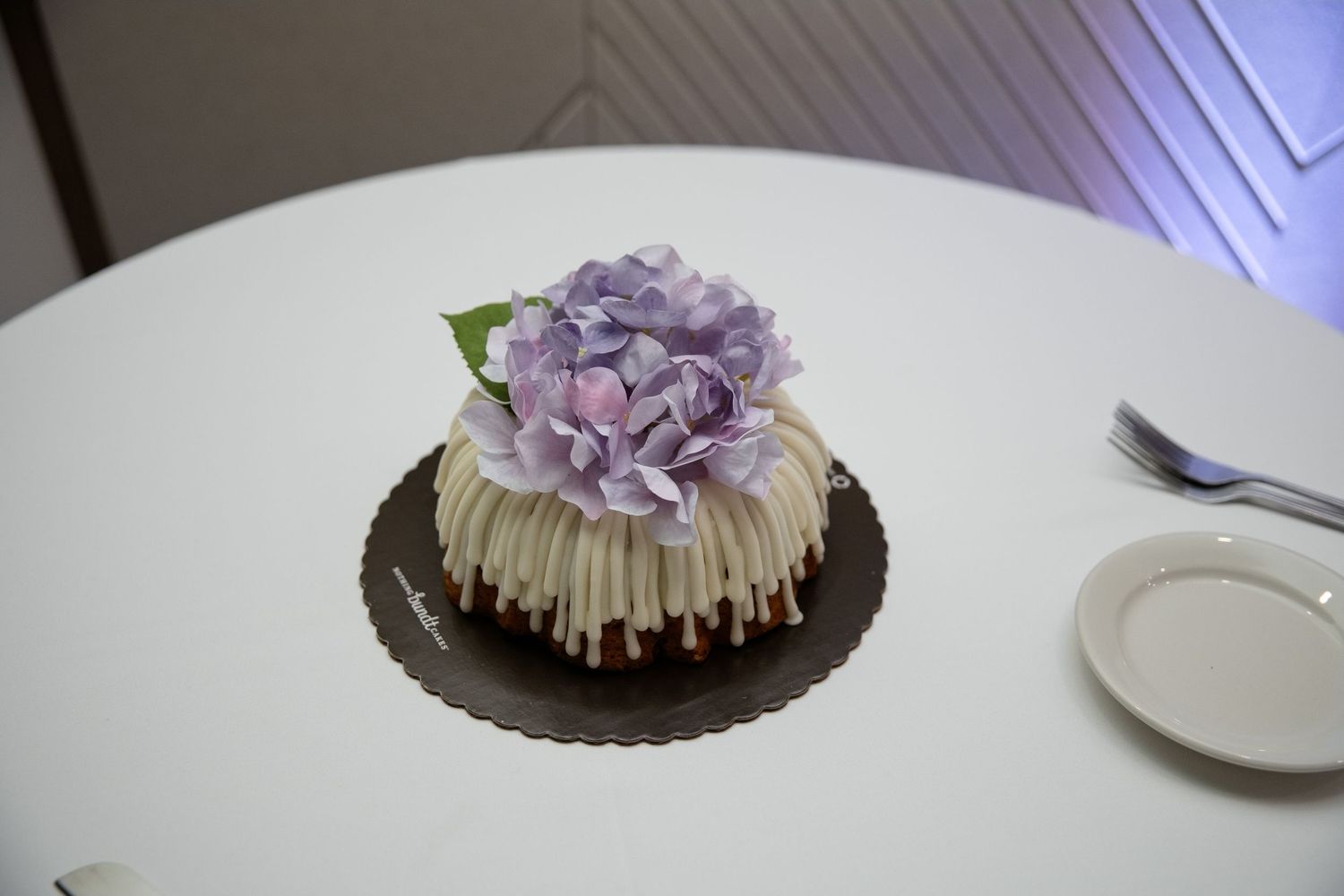 Elegant white bundt cake topped with purple hydrangea flowers on a brown cake board on a white table.