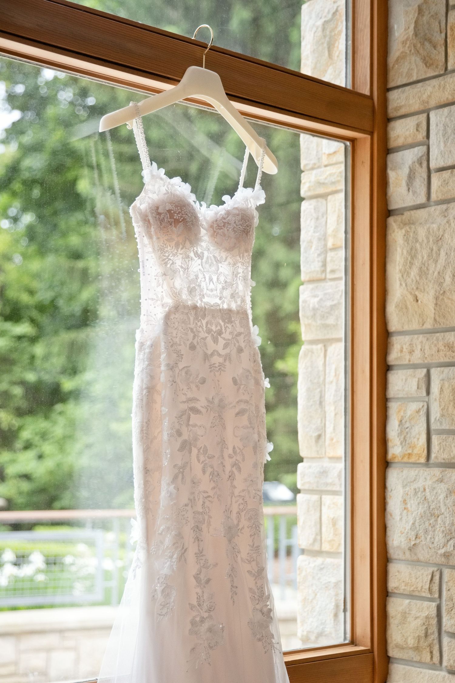 Elegant lace wedding dress hanging on a wooden hanger in a window with stone wall backdrop.