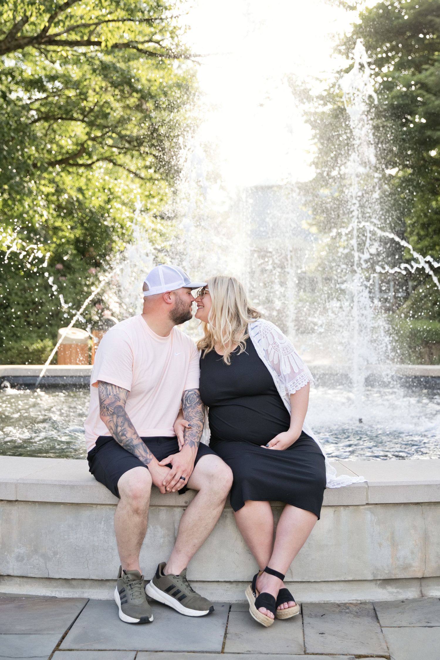 Couple sharing a kiss while sitting on fountain edge, woman in black maternity dress, man with tattoos.