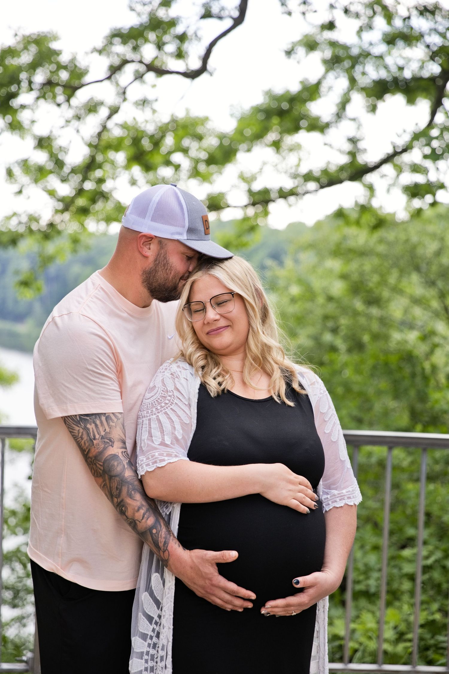 Expectant couple shares tender moment outdoors, man embraces pregnant woman from behind near scenic overlook.