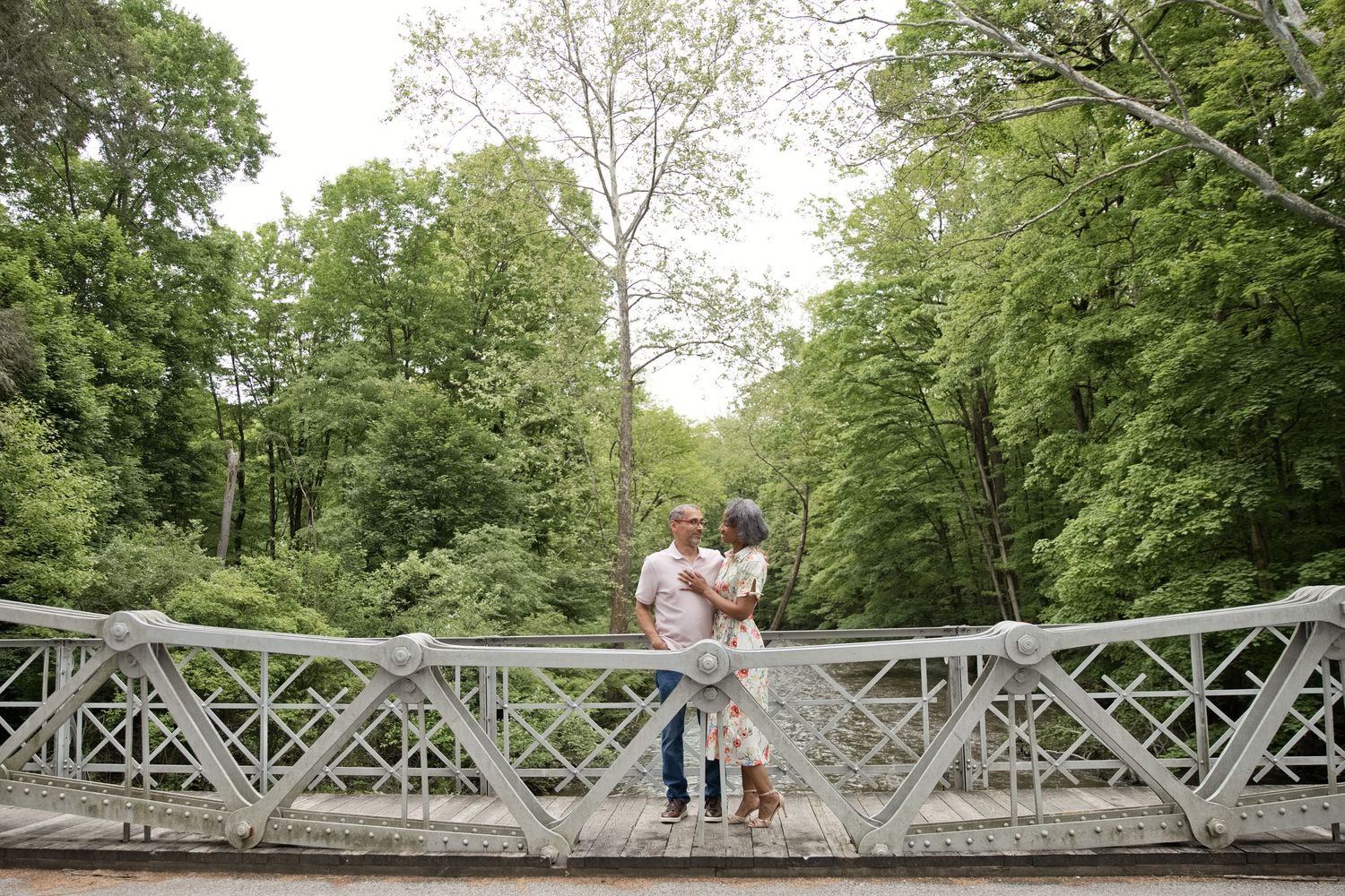 Couple embracing on a white ornate bridge surrounded by vibrant green trees.