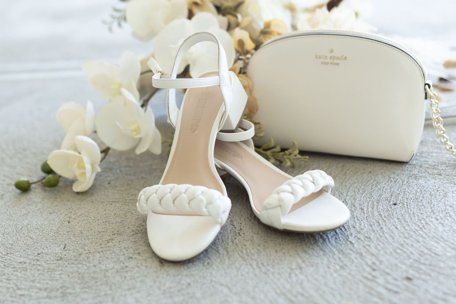 Elegant white bridal sandals with braided details displayed next to a matching clutch and flowers.