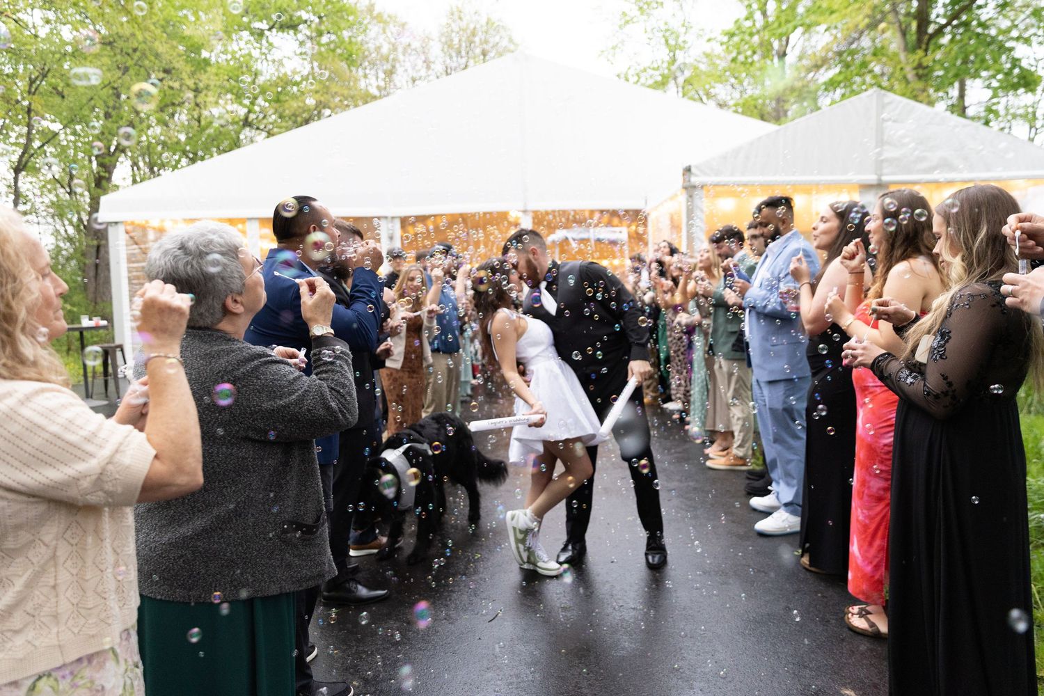 Newlyweds walk through a joyful bubble send-off surrounded by guests outside a white wedding tent.