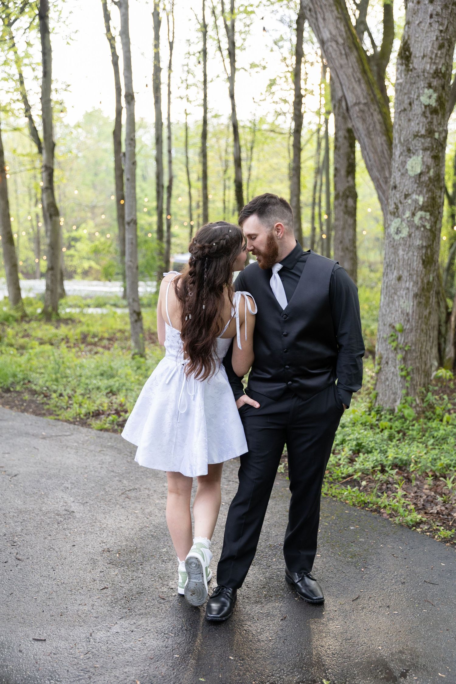 A bride in a white dress and groom in a black suit share a romantic moment on a tree-lined path.