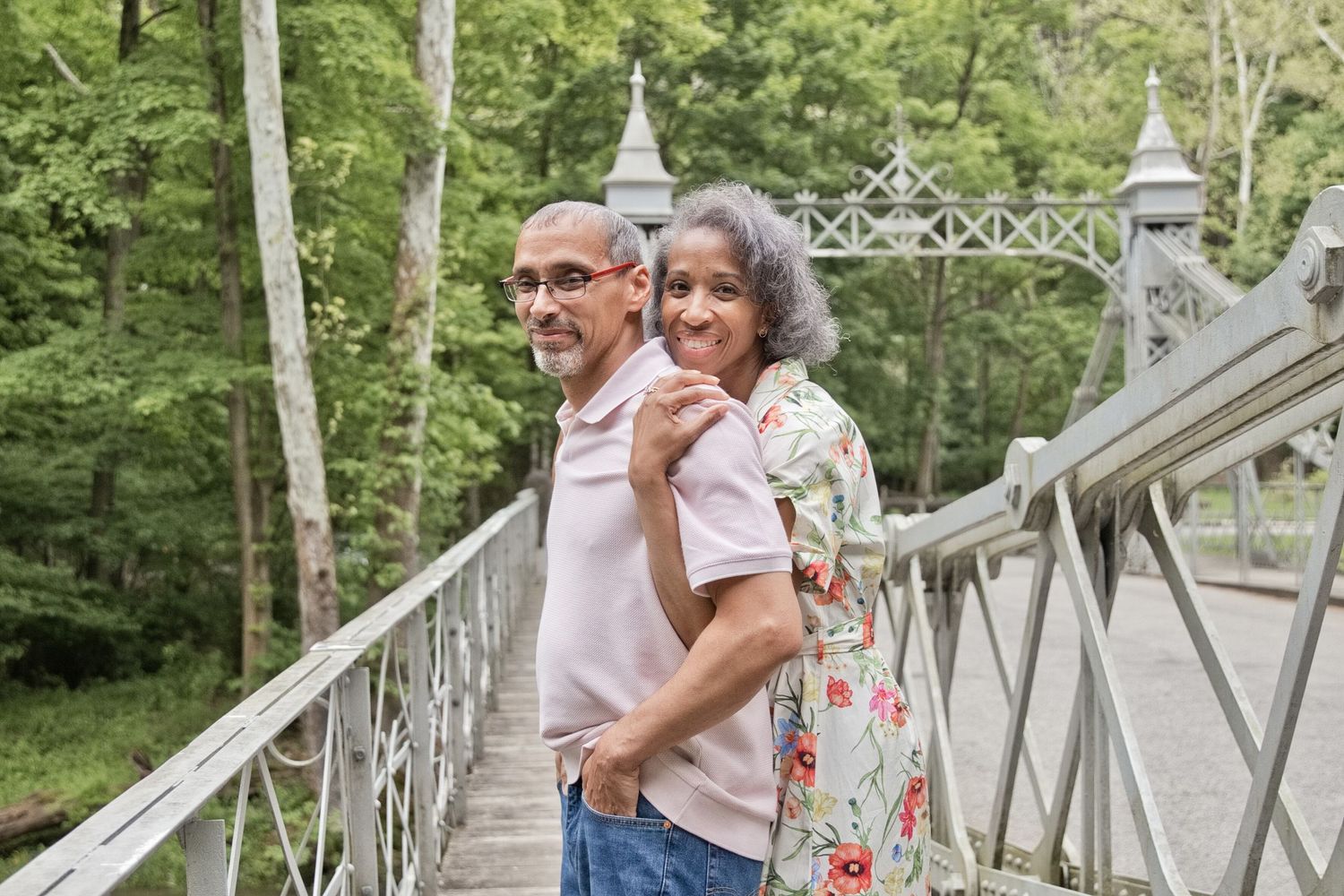 An older couple smiles together on a white bridge surrounded by lush green trees.