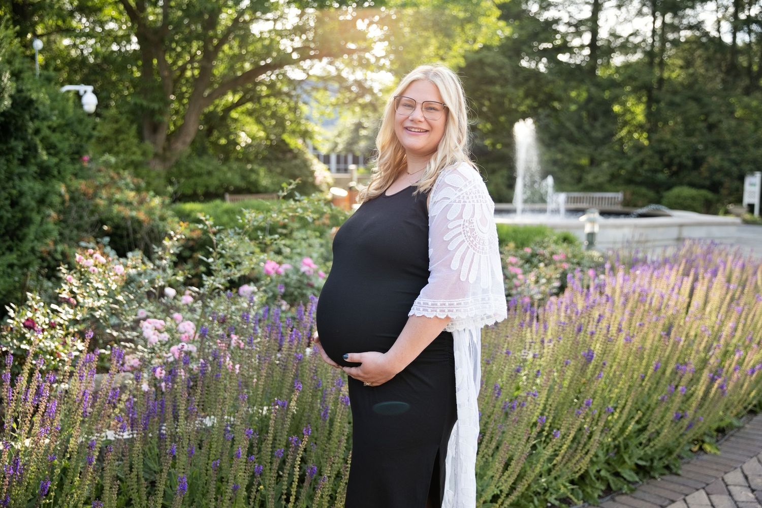 Pregnant woman in black dress and white cardigan smiling in a garden with lavender and pink flowers.