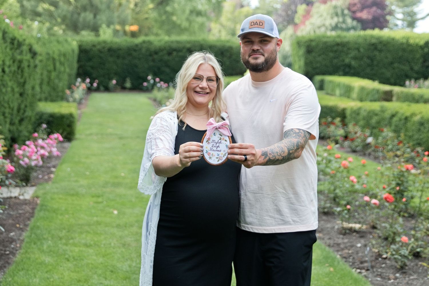 Pregnant couple posing in a rose garden, holding an ultrasound photo, surrounded by lush green hedges.