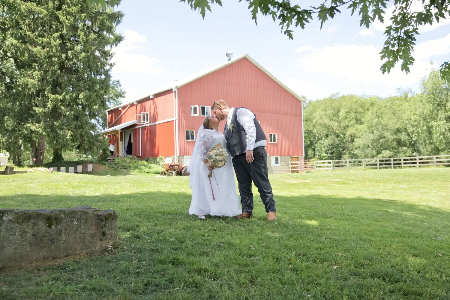 Bride and groom share a kiss on green lawn before a red barn on their rustic outdoor wedding day.
