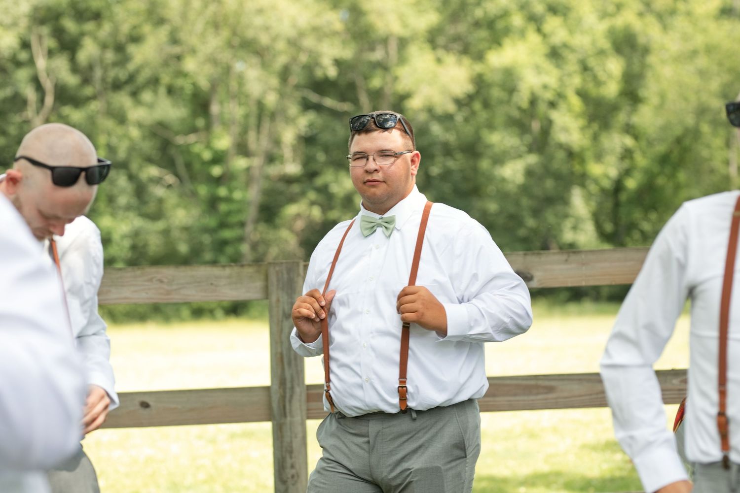 Groomsman in white shirt, green bow tie, and brown suspenders stands outdoors near a wooden fence at a wedding.