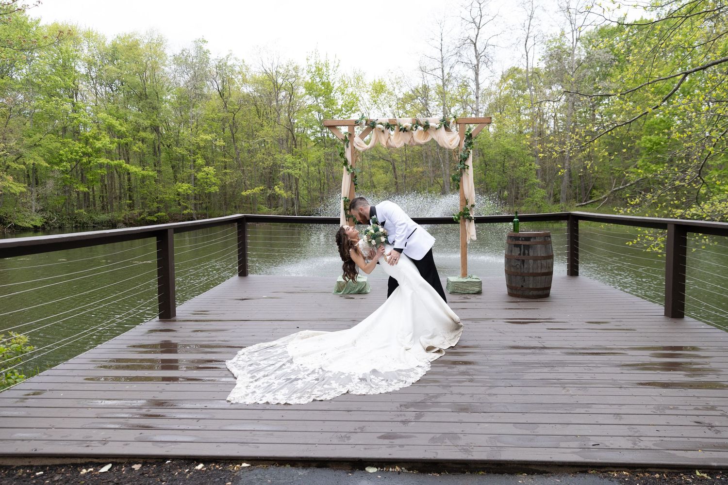 Bride and groom share a romantic dip kiss on a wooden deck overlooking a serene lake surrounded by lush green trees.