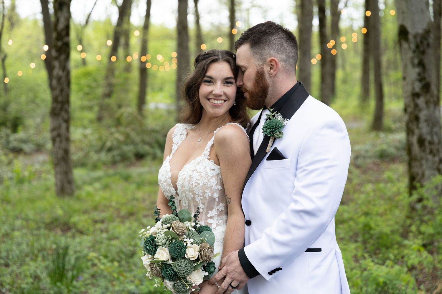Bride and groom share a romantic moment in a woodland setting, bride holding white bouquet, groom in white tuxedo jacket.