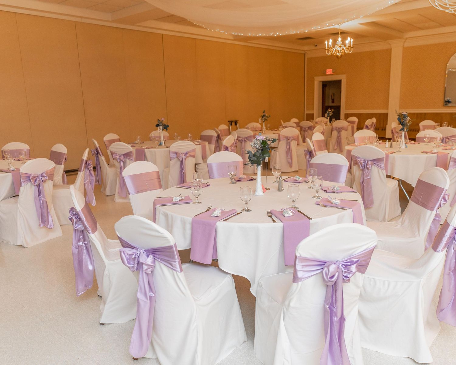 Elegant banquet hall with round tables, white chair covers, and lavender sashes for a wedding reception.