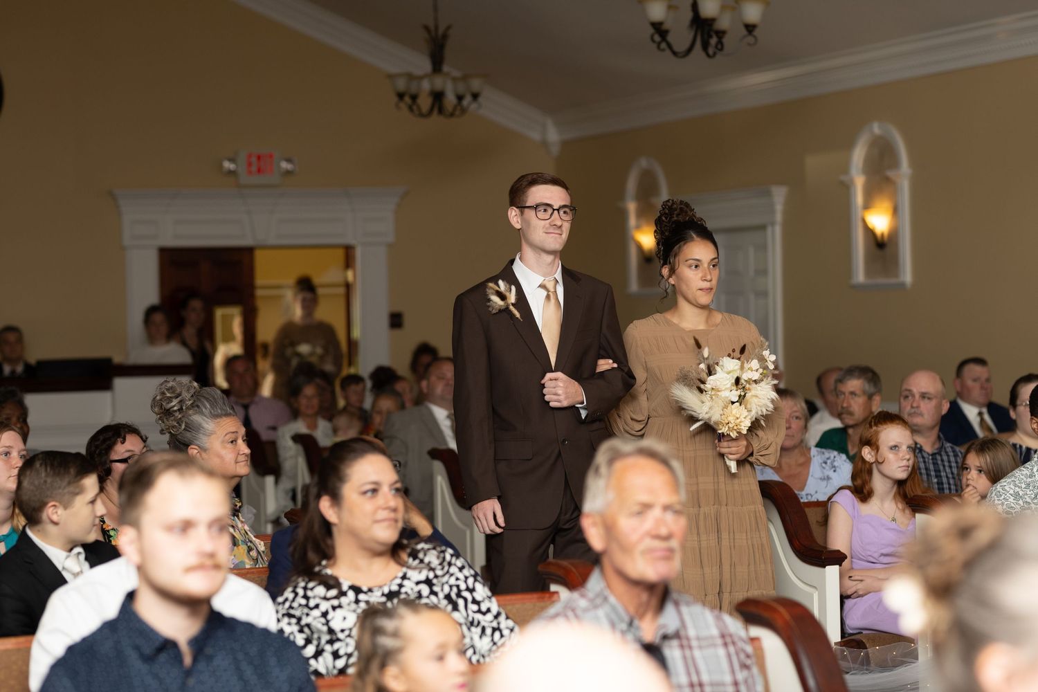 Guests seated in a formal venue during a wedding ceremony with chandelier lighting overhead.