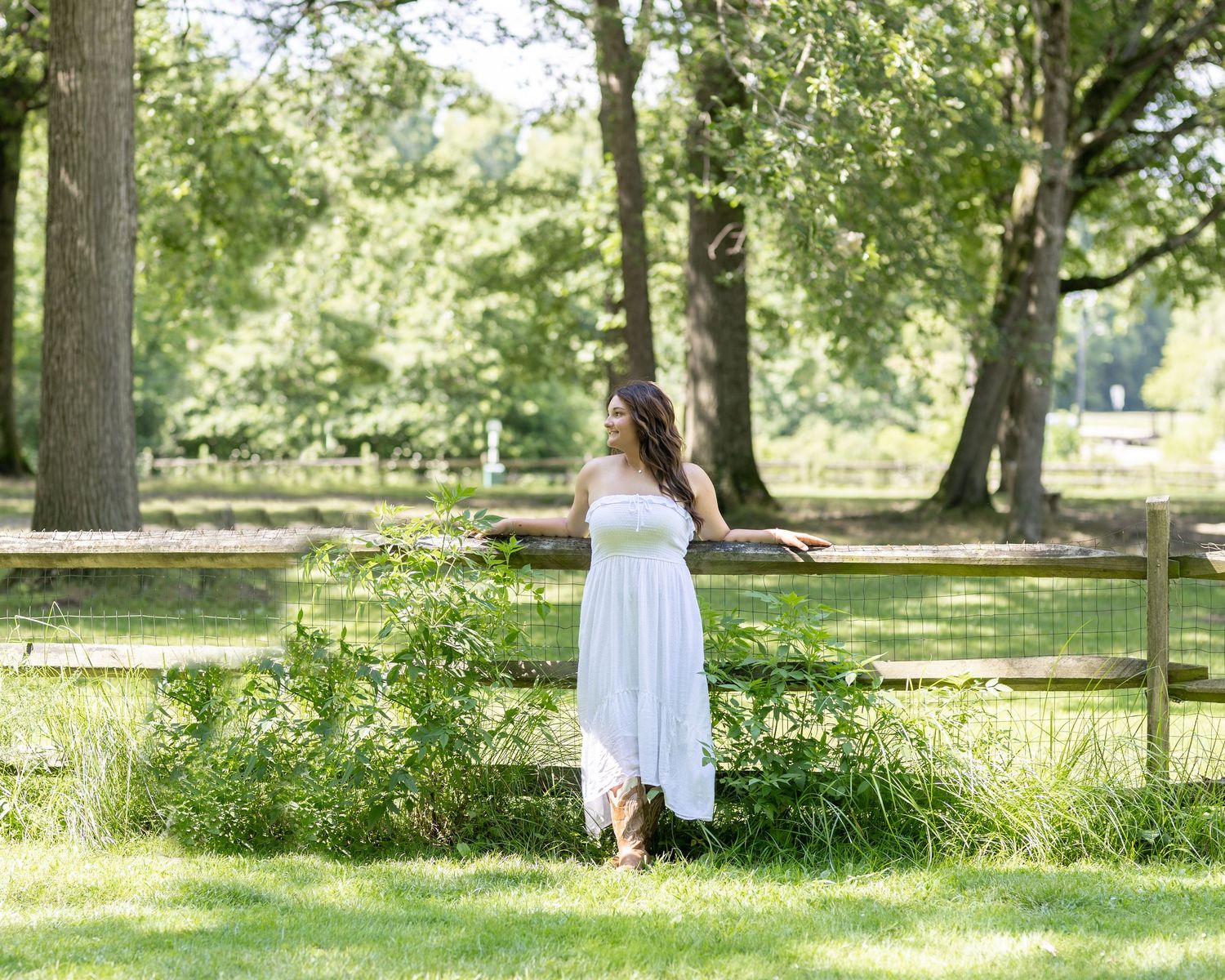 A white sundress flows in a sunny garden setting with lush trees and a wooden fence in the background.