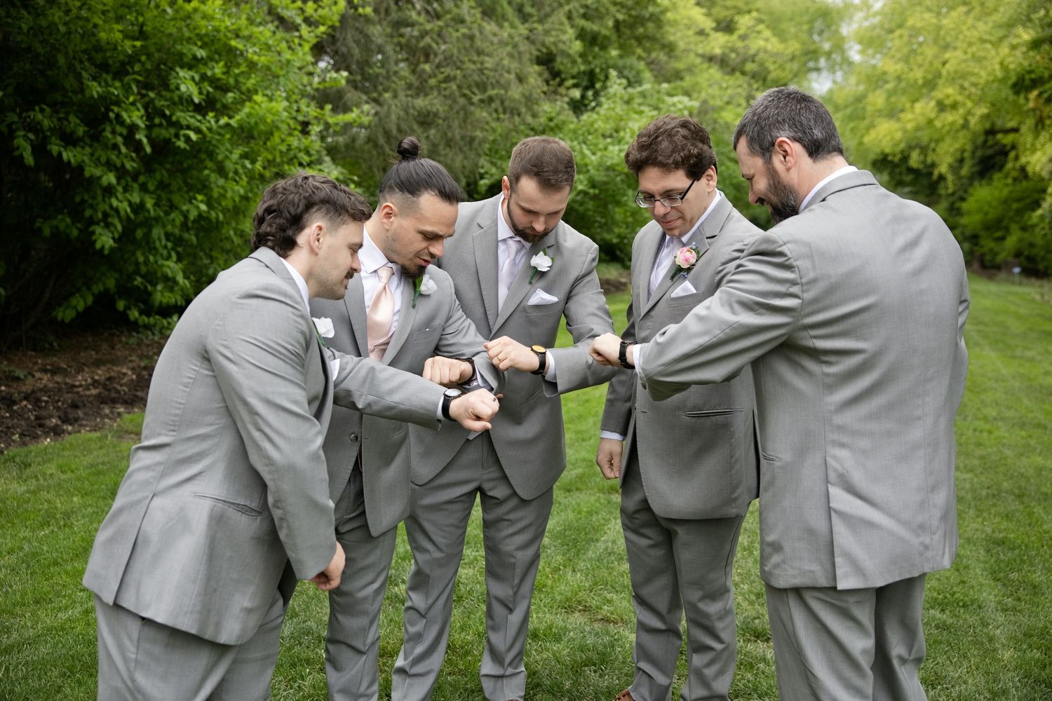 Groomsmen in gray suits huddle together admiring watches outdoors on green lawn.