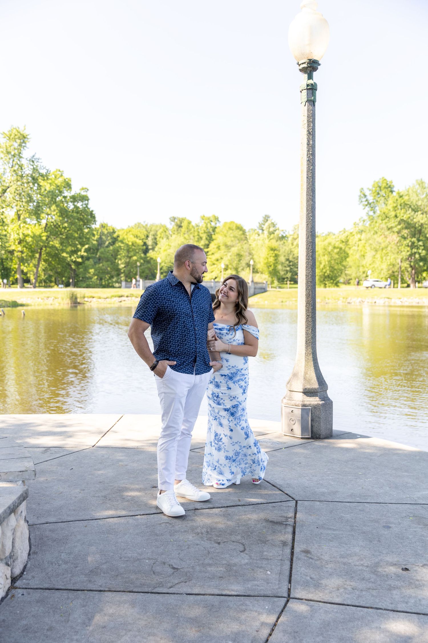 Couple standing together by a lake near a tall lamppost in a sunny green park.