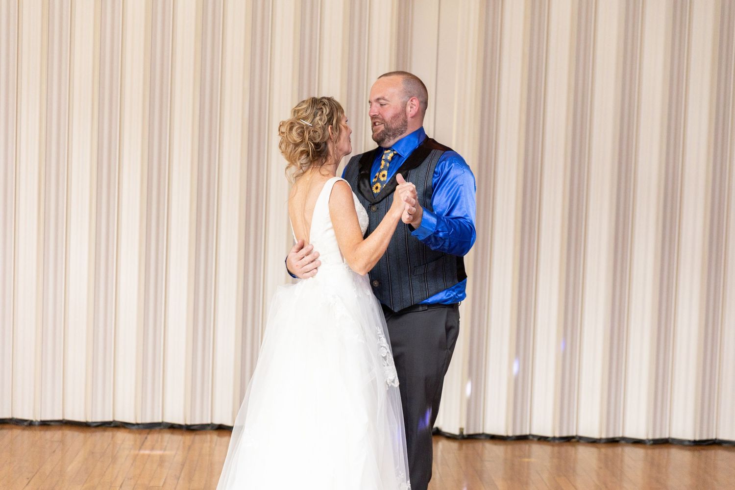 Bride and groom share their first dance at wedding reception, groom wearing royal blue shirt and black vest.