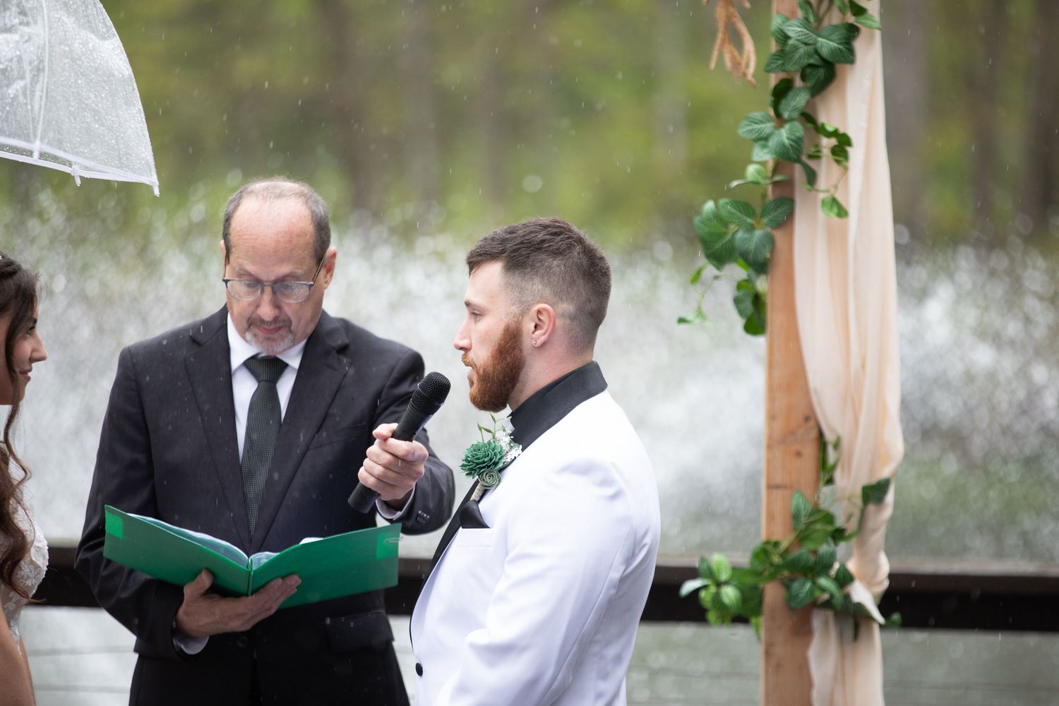 Groom reciting wedding vows at outdoor ceremony with officiant holding green folder, ivy-adorned arch in background.