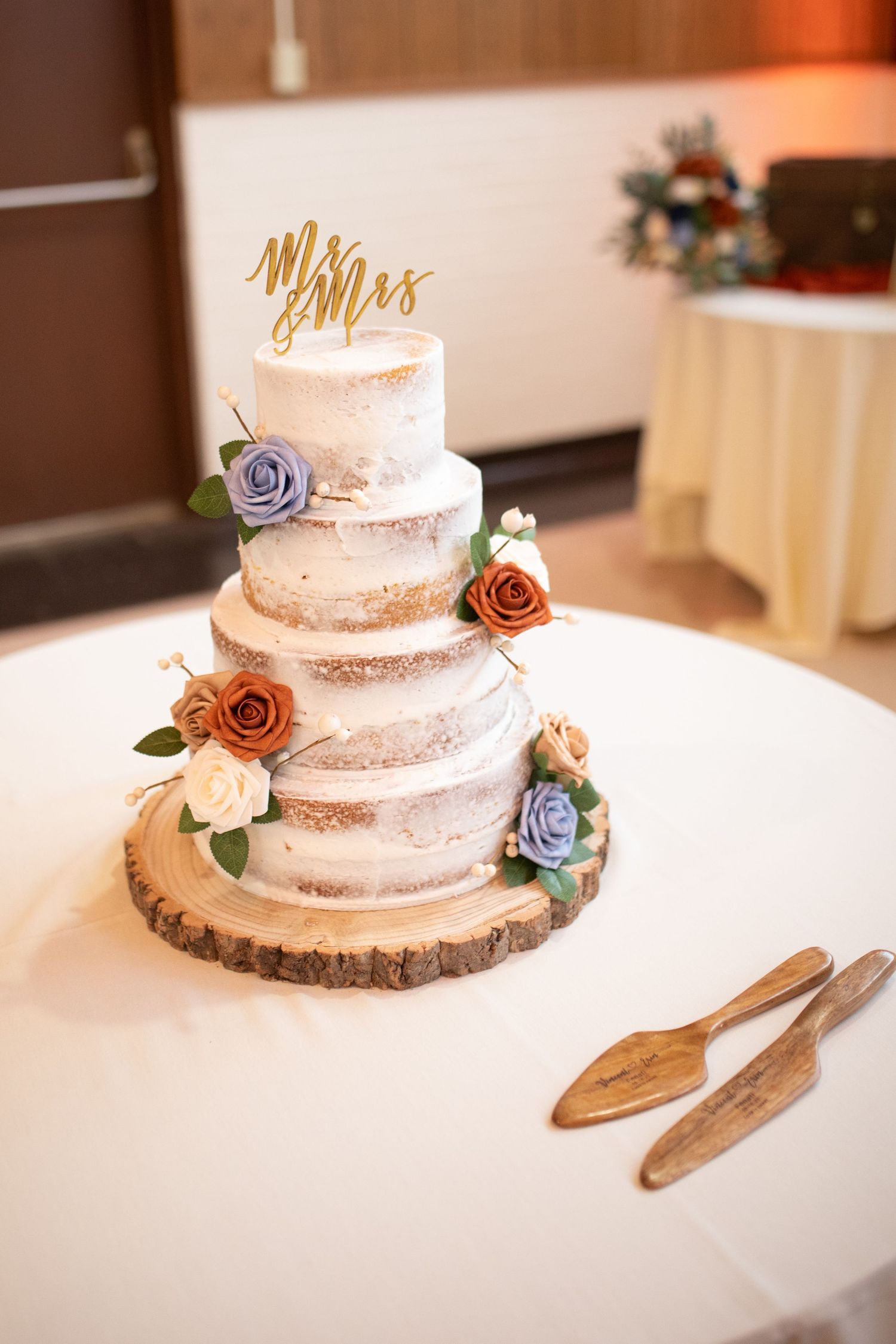 A semi-naked wedding cake with purple and rust colored sugar flowers on a wooden cake stand.