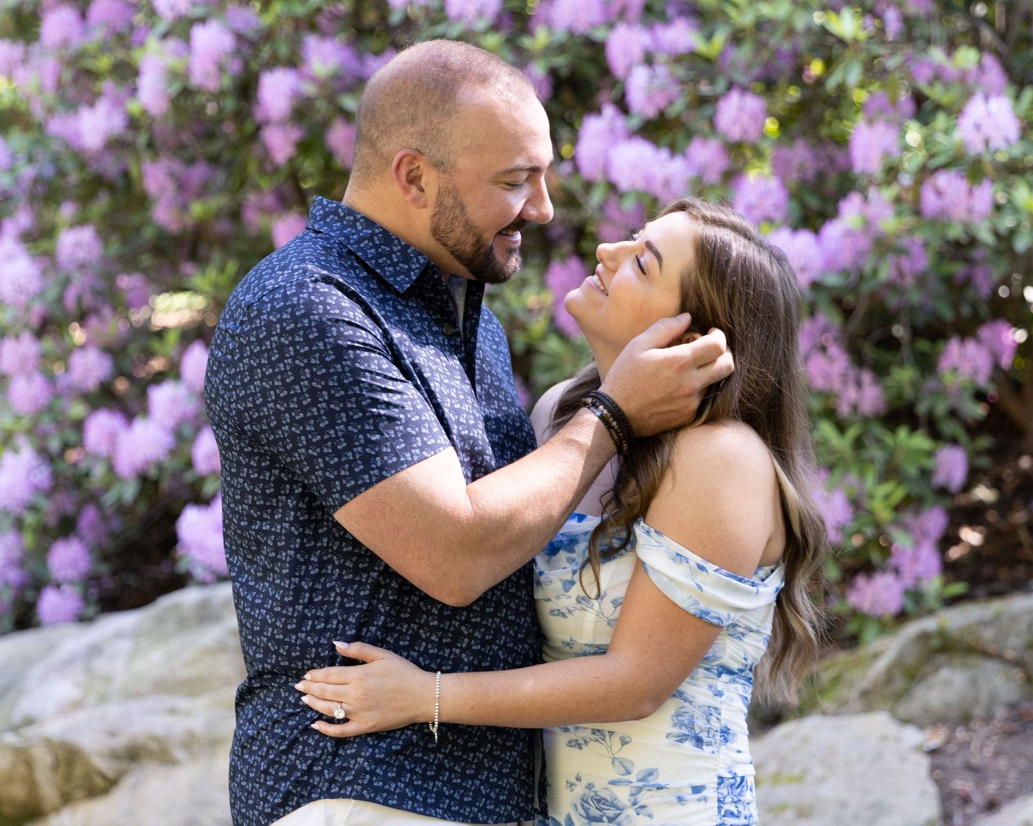 Couple embracing romantically in front of blooming purple rhododendron bushes in a lush garden setting.