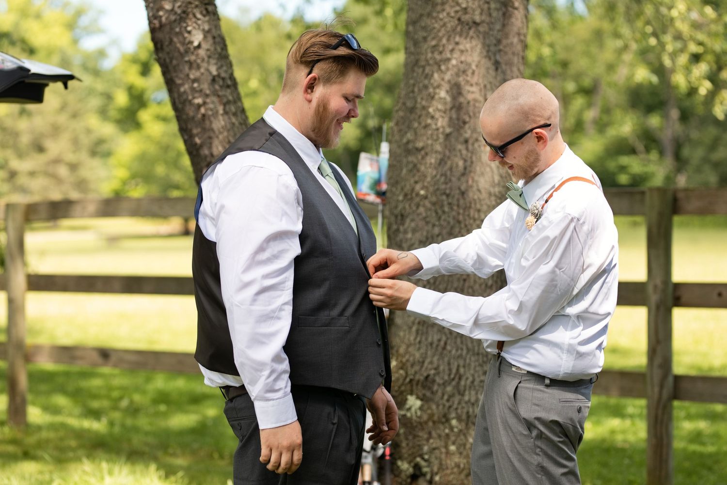 Groom in vest being helped by groomsman with boutonniere outdoors at rustic wedding ceremony.