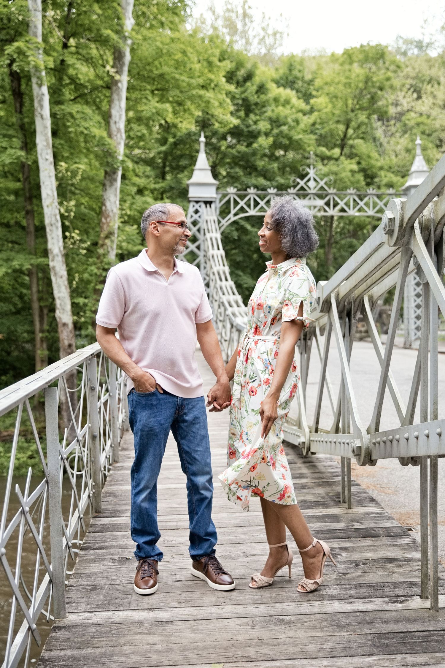 A couple holds hands on a decorative bridge surrounded by lush green trees, smiling at each other.