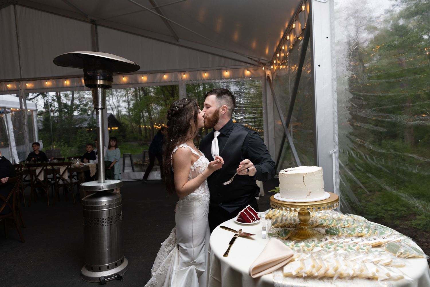 Bride and groom share a kiss near their wedding cake under a tented reception venue with string lights.