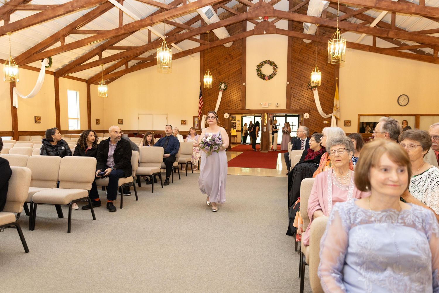 A bridesmaid walks down the aisle of a decorated church during a wedding ceremony with seated guests.