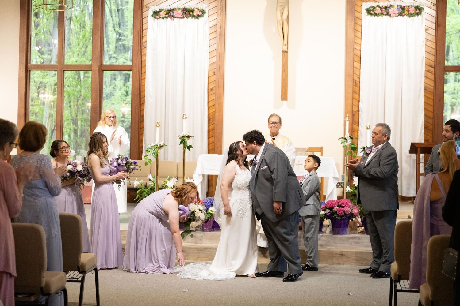 Bride and groom share first kiss at church altar surrounded by bridesmaids in lavender dresses and groomsmen during wedding ceremony.