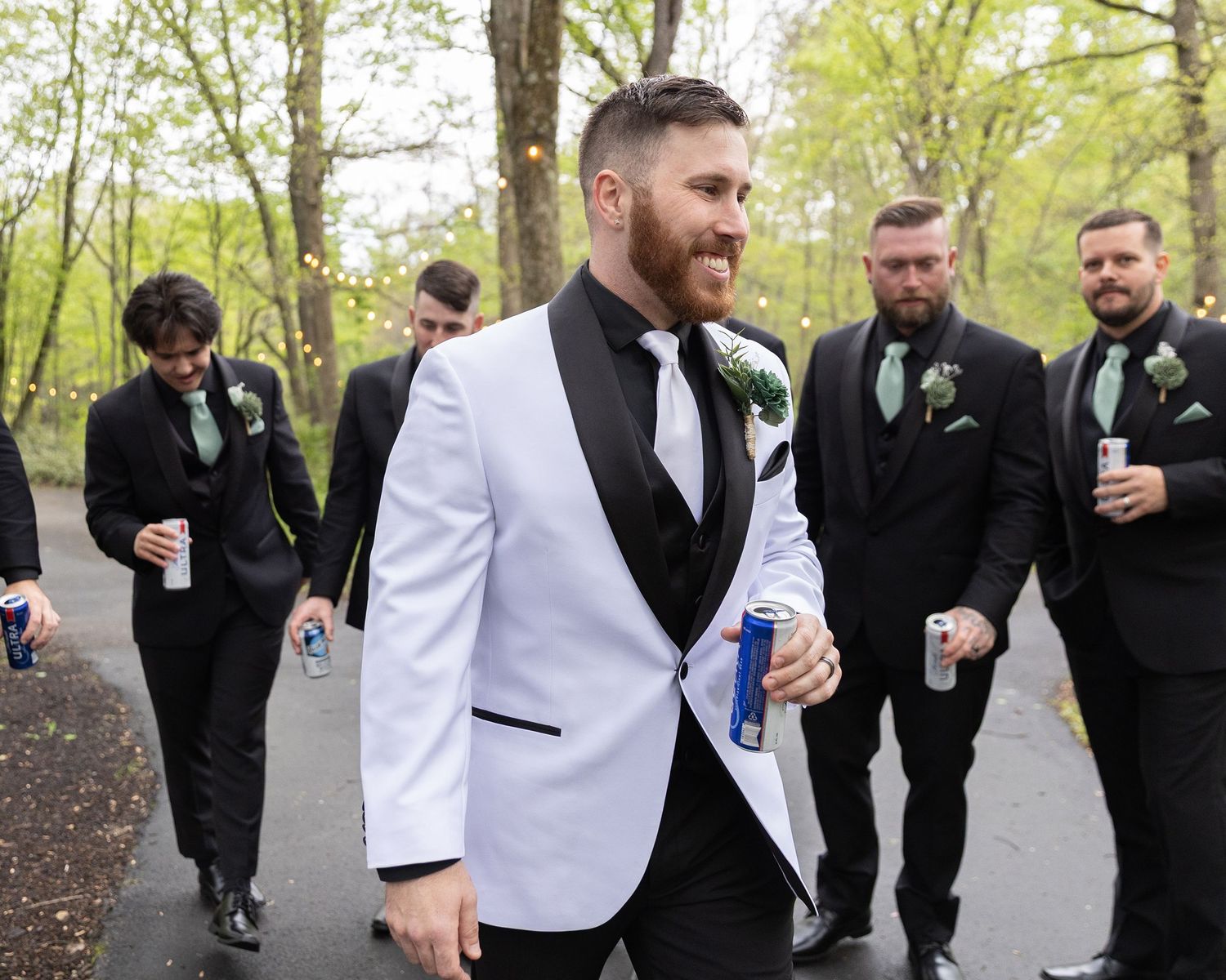 Groom in white tuxedo jacket walks with groomsmen in black suits and green ties on a tree-lined path.